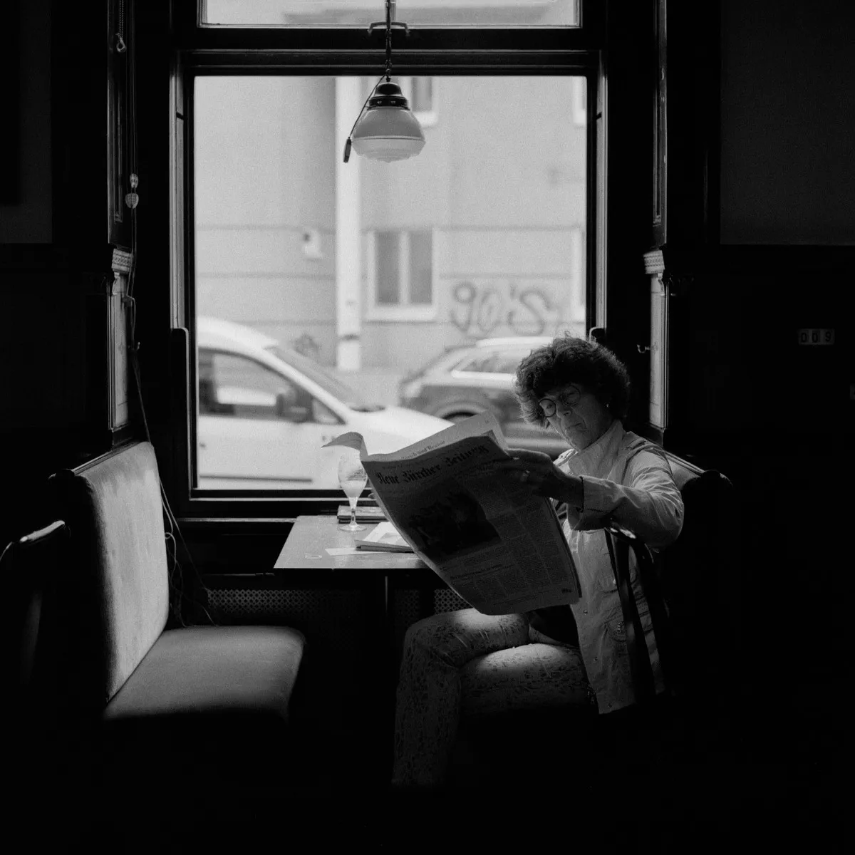A woman reads a newspaper at a window table in a Viennese café, her face visible above the raised broadsheet, a pendant lamp overhead and parked cars on the street outside
