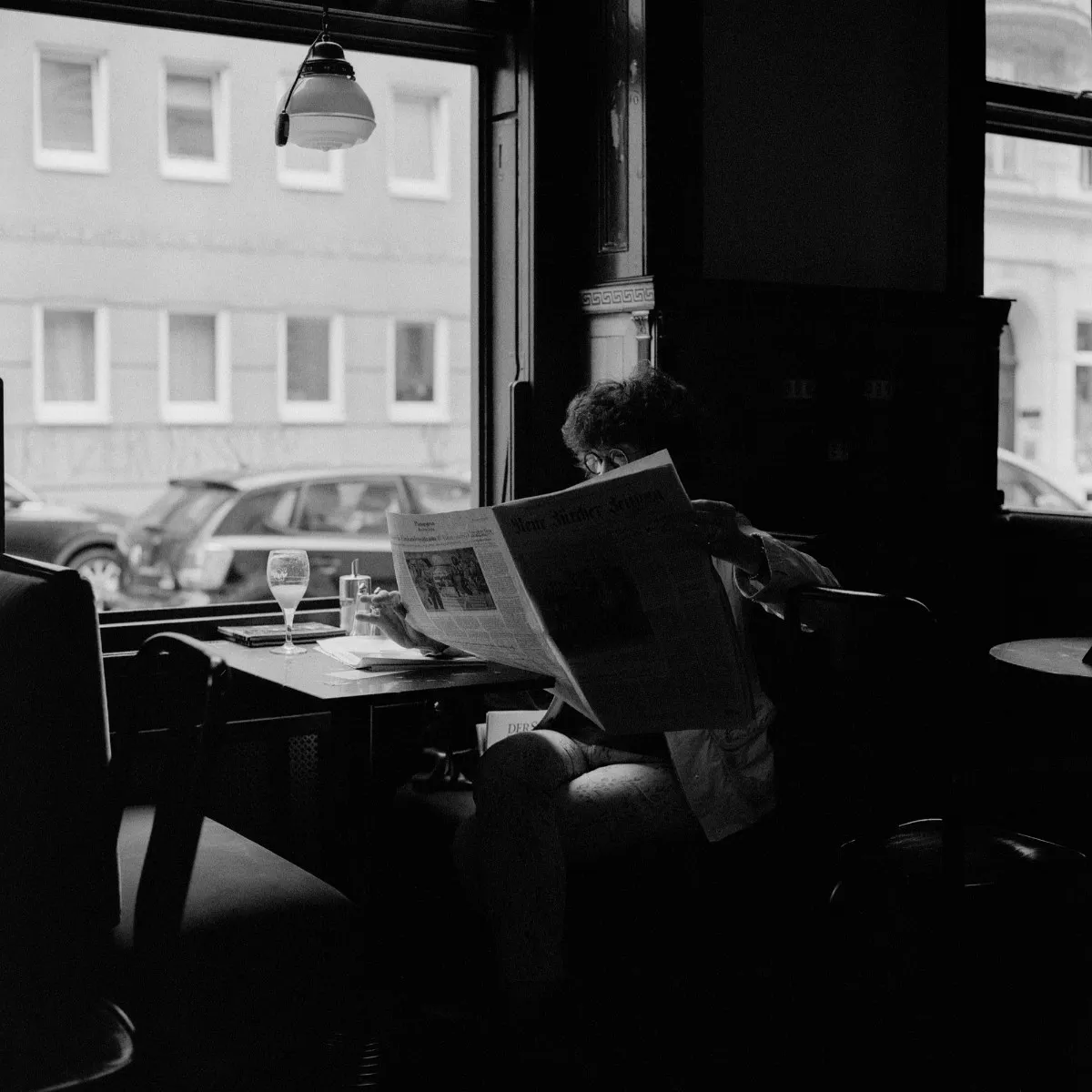 A wider view of a Viennese café window table, the reader's face hidden behind a raised newspaper, marble-topped tables nearby and parked cars and a pale façade beyond the glass